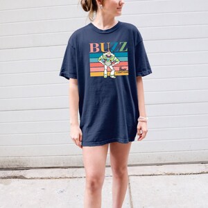May include: A navy blue t-shirt with a retro-style graphic of Buzz Lightyear from Toy Story. The graphic features Buzz in his space ranger suit with a colorful striped background and the text "Buzz Lightyear".