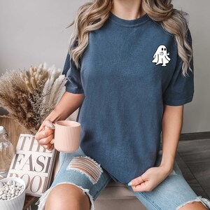 May include: A woman wearing a dark blue t-shirt with a white ghost graphic. The ghost is waving. The woman is holding a pink mug.