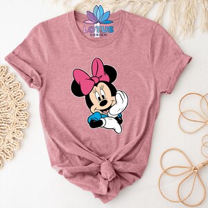 K&ouml;nnte beinhalten: Ein pinkfarbenes T-Shirt mit einem Cartoon-Bild von Minnie Mouse. Das Bild zeigt Minnie Mouse mit einer rosa Schleife und einem blauen Kleid. Der Text "LOTUS DESIGN" ist auf dem Shirt aufgedruckt.