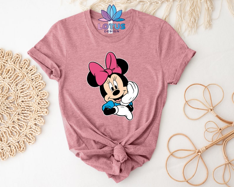 K&ouml;nnte beinhalten: Ein pinkfarbenes T-Shirt mit einem Cartoon-Bild von Minnie Mouse. Das Bild zeigt Minnie Mouse mit einer rosa Schleife und einem blauen Kleid. Der Text "LOTUS DESIGN" ist auf dem Shirt aufgedruckt.