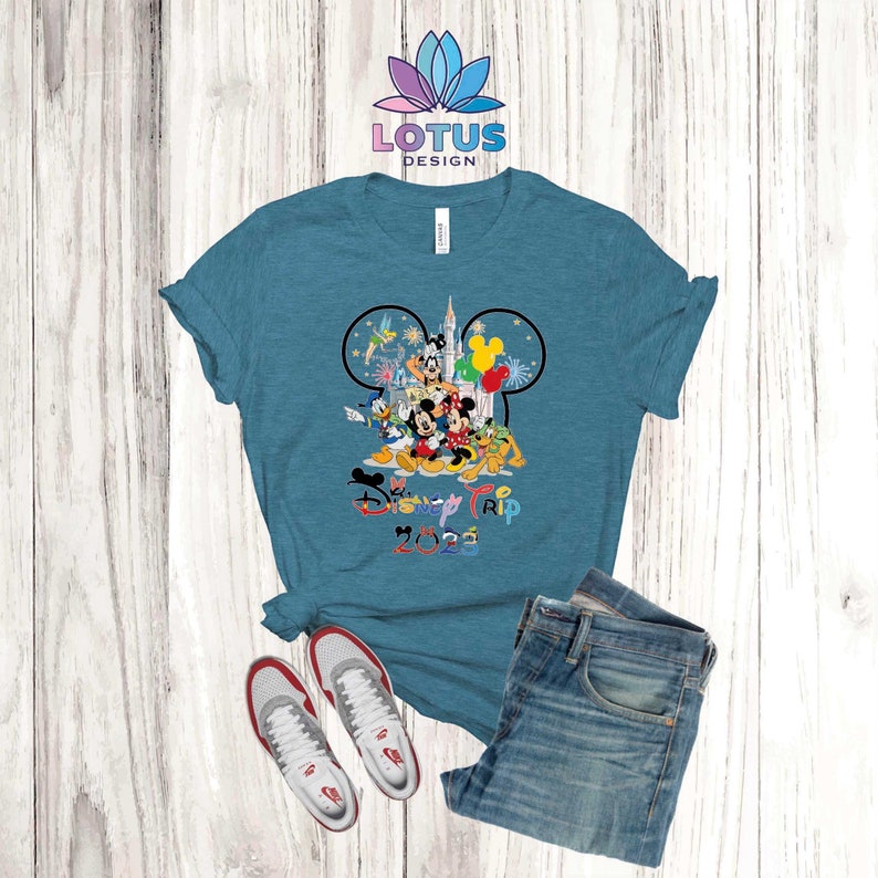 Puede incluir: Una camiseta azul turquesa con un gr&aacute;fico de Mickey Mouse y otros personajes de Disney frente al Castillo de Cenicienta. El texto "Disney Trip 2023" est&aacute; impreso debajo del gr&aacute;fico.