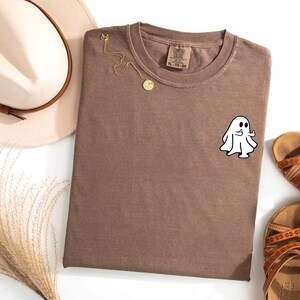 May include: A brown t-shirt with a white ghost giving a thumbs up. The ghost is on the left side of the shirt.