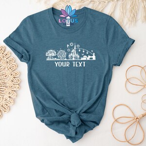K&ouml;nnte beinhalten: Ein blaues T-Shirt mit einem wei&szlig;en Umriss der Disney World Skyline, einschlie&szlig;lich Cinderella's Castle, Spaceship Earth und dem Haunted Mansion. Der Text "YOUR TEXT" ist unterhalb der Skyline gedruckt.