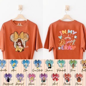 Puede incluir: Camiseta naranja con un gr&aacute;fico de Bella de La Bella y la Bestia en una silueta de oreja de Mickey Mouse. La parte posterior de la camiseta tiene el texto "In My Disney Era" con un gr&aacute;fico colorido del castillo de Disney. La camiseta est&aacute; disponible en 12 dise&ntilde;os diferentes de princesas Disney.