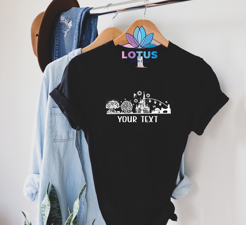 K&ouml;nnte beinhalten: Schwarzes T-Shirt mit einer wei&szlig;en Silhouette einer Themenpark-Skyline, einschlie&szlig;lich eines Schlosses, eines Riesenrads und eines Baumes. Der Text "YOUR TEXT" befindet sich unterhalb der Skyline.