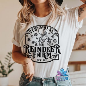 May include: White t-shirt with a black graphic design featuring a reindeer and snowman with the text "Sven & Olaf's Reindeer Farm Est. 2013".