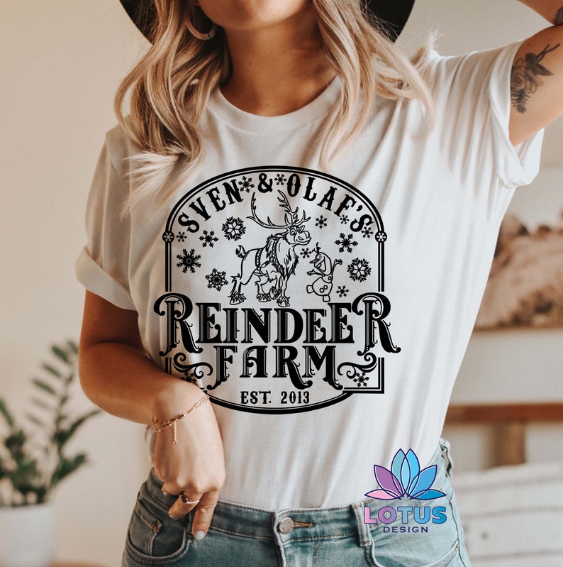 May include: White t-shirt with a black graphic design featuring a reindeer and snowman with the text "Sven & Olaf's Reindeer Farm Est. 2013".
