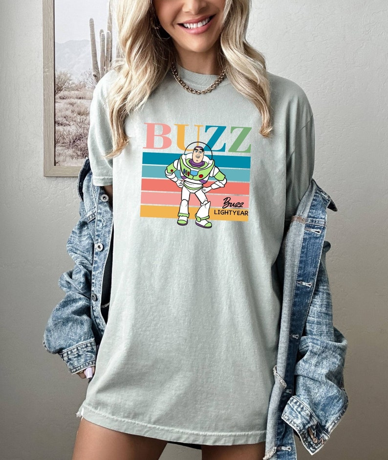 May include: A light green t-shirt with a retro-style graphic of Buzz Lightyear from Toy Story. The graphic features Buzz Lightyear standing with his arms crossed, with the text "Buzz" above him and "Buzz Lightyear" below him.