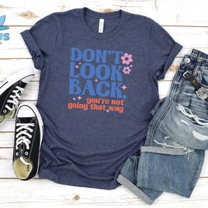May include: A navy blue t-shirt with the text "Don't Look Back. You're not going that way" in blue, pink, and orange lettering. The shirt is styled with a pair of black sneakers and blue jeans.