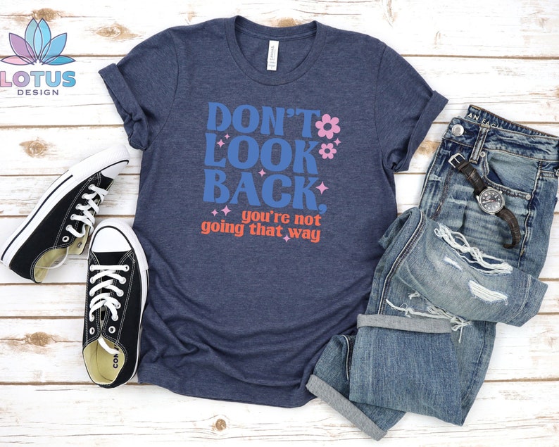 May include: A navy blue t-shirt with the text "Don't Look Back. You're not going that way" in blue, pink, and orange lettering. The shirt is styled with a pair of black sneakers and blue jeans.