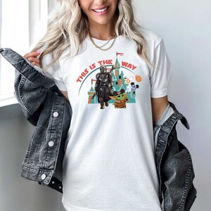 May include: White t-shirt featuring a graphic of a castle, a Mandalorian, and Grogu. The text "This is the way" is printed in a circle around the castle.