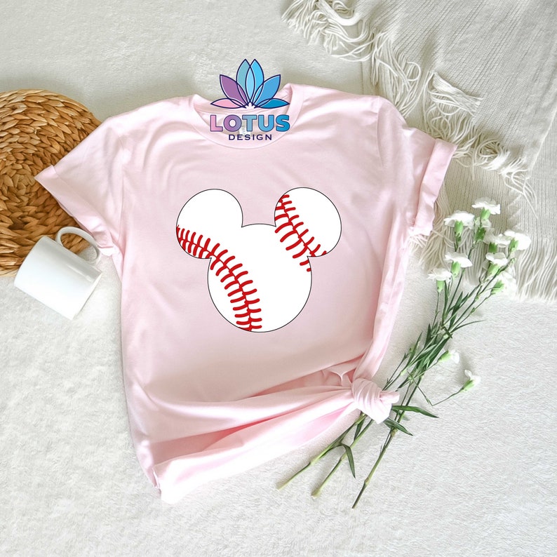 May include: Pink t-shirt with a white baseball shaped like Mickey Mouse's head. The baseball has red stitching.