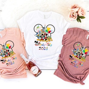 Puede incluir: Tres camisetas con un dise&ntilde;o de oreja de Mickey Mouse con personajes de Disney y el texto "Disney Trip 2023". Las camisetas son de color rosa, blanco y marr&oacute;n.