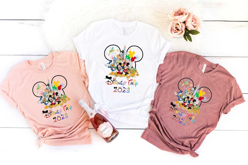 Puede incluir: Tres camisetas con un dise&ntilde;o de oreja de Mickey Mouse con personajes de Disney y el texto "Disney Trip 2023". Las camisetas son de color rosa, blanco y marr&oacute;n.