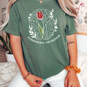 May include: A green t-shirt featuring a white outline of a brain with a red tulip growing from it. The text "Parkinson's Awareness" is printed below the image.