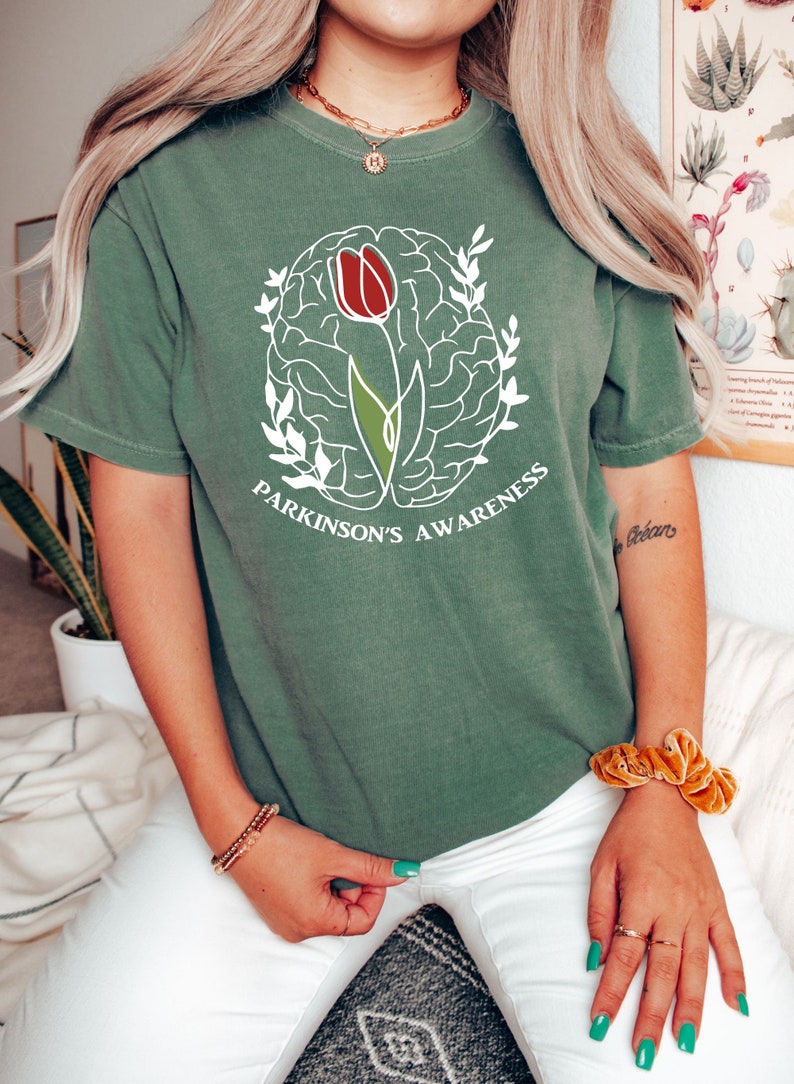 May include: A green t-shirt featuring a white outline of a brain with a red tulip growing from it. The text "Parkinson's Awareness" is printed below the image.