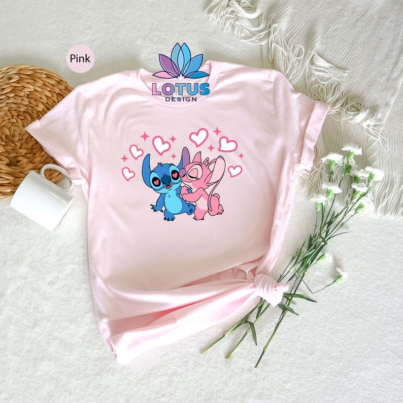K&ouml;nnte beinhalten: Pinkes T-Shirt mit einem Motiv von Stitch und Angel aus Lilo & Stitch. Das Motiv zeigt Stitch und Angel, die sich umarmen, mit Herzen und Glitzer um sie herum. Der Text "LOTUS DESIGN" ist auf das T-Shirt gedruckt.