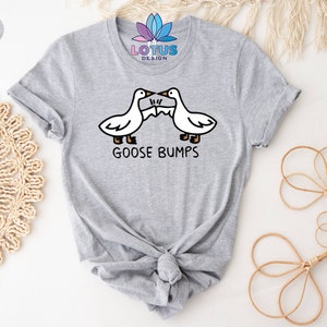 Op de afbeelding: Een grijs T-shirt met een afbeelding van twee ganzen die tegen elkaar aanbotsen. De tekst "GOOSE BUMPS" staat onder de ganzen.