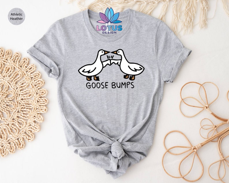 Op de afbeelding: Een grijs T-shirt met een afbeelding van twee ganzen die tegen elkaar aanbotsen. De tekst "GOOSE BUMPS" staat onder de ganzen.