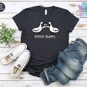 Op de afbeelding: Donkergrijs t-shirt met een afbeelding van twee ganzen die elkaar aankijken met de tekst "Goose Bumps". Het shirt ligt plat op een wit houten oppervlak met een paar witte canvas schoenen en een blauwe jeans.