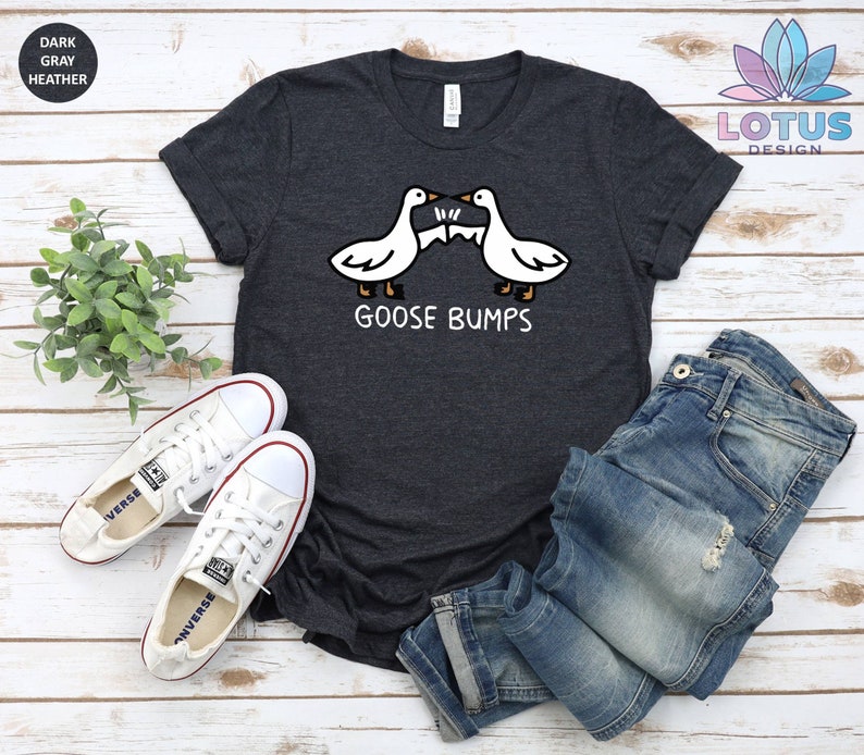 Op de afbeelding: Donkergrijs t-shirt met een afbeelding van twee ganzen die elkaar aankijken met de tekst "Goose Bumps". Het shirt ligt plat op een wit houten oppervlak met een paar witte canvas schoenen en een blauwe jeans.