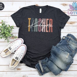 K&ouml;nnte beinhalten: Dunkelgraues T-Shirt mit dem Text "Kindergarten Teacher" in bunten Streifen. Das Shirt ist auf einem wei&szlig;en Holzgrund mit einem Paar wei&szlig;en Turnschuhen und einer blauen Jeans zu sehen.
