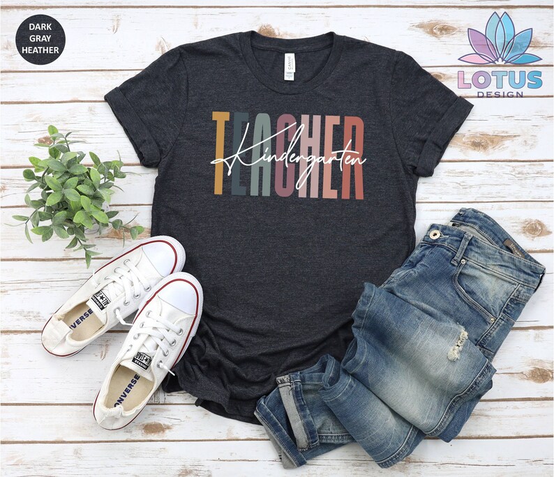 K&ouml;nnte beinhalten: Dunkelgraues T-Shirt mit dem Text "Kindergarten Teacher" in bunten Streifen. Das Shirt ist auf einem wei&szlig;en Holzgrund mit einem Paar wei&szlig;en Turnschuhen und einer blauen Jeans zu sehen.