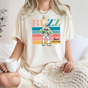May include: A white t-shirt with a retro-style graphic of Buzz Lightyear from Toy Story. The graphic features a colorful striped background with the text "Buzz" in pink, blue, and green letters. The text "Buzz Lightyear" is written in smaller letters below the graphic.