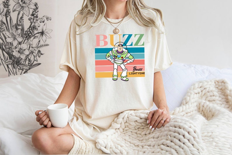 May include: A white t-shirt with a retro-style graphic of Buzz Lightyear from Toy Story. The graphic features a colorful striped background with the text "Buzz" in pink, blue, and green letters. The text "Buzz Lightyear" is written in smaller letters below the graphic.