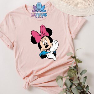 K&ouml;nnte beinhalten: Ein rosa T-Shirt mit einem Aufdruck von Minnie Maus, die eine rosa Schleife tr&auml;gt. Der Text "LOTUS DESIGN" ist auf dem Shirt gedruckt.