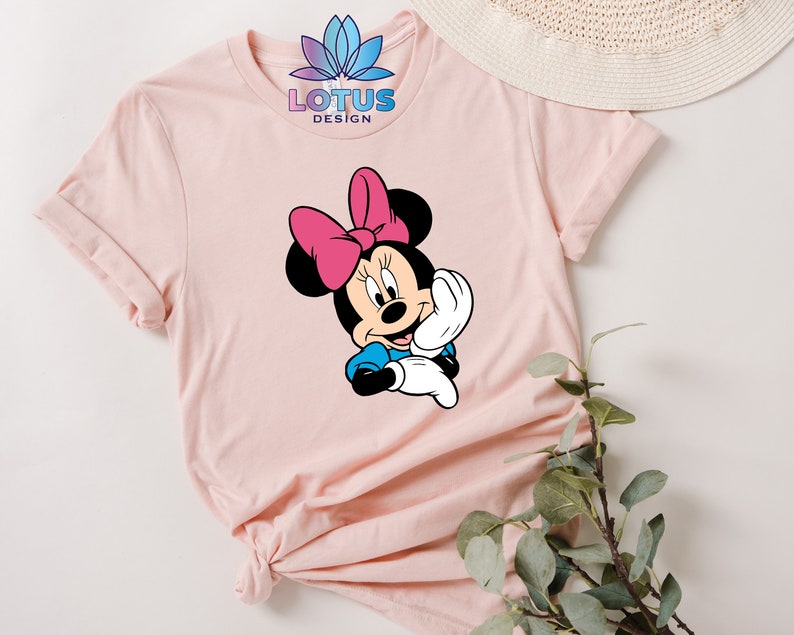 K&ouml;nnte beinhalten: Ein rosa T-Shirt mit einem Aufdruck von Minnie Maus, die eine rosa Schleife tr&auml;gt. Der Text "LOTUS DESIGN" ist auf dem Shirt gedruckt.