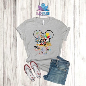 Puede incluir: Una camiseta gris con un contorno negro de oreja de Mickey Mouse. Dentro del contorno hay varios personajes de Disney, incluidos Mickey Mouse, Minnie Mouse, Donald Duck, Goofy y Pluto. El texto "Disney Trip 2023" est&aacute; impreso debajo de los personajes en colores arco&iacute;ris.