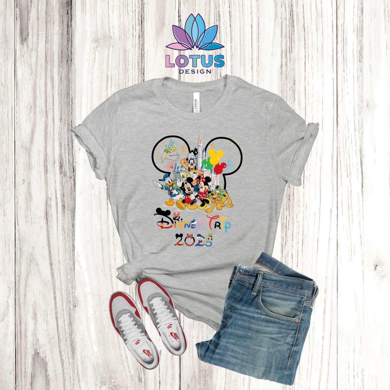 Puede incluir: Una camiseta gris con un contorno negro de oreja de Mickey Mouse. Dentro del contorno hay varios personajes de Disney, incluidos Mickey Mouse, Minnie Mouse, Donald Duck, Goofy y Pluto. El texto "Disney Trip 2023" est&aacute; impreso debajo de los personajes en colores arco&iacute;ris.