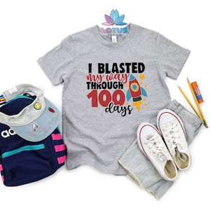 May include: A gray t-shirt with the text "I blasted my way through 100 days" and a red rocket ship graphic. The shirt is laid out on a white background with a pair of white sneakers, a denim skirt, and a backpack.