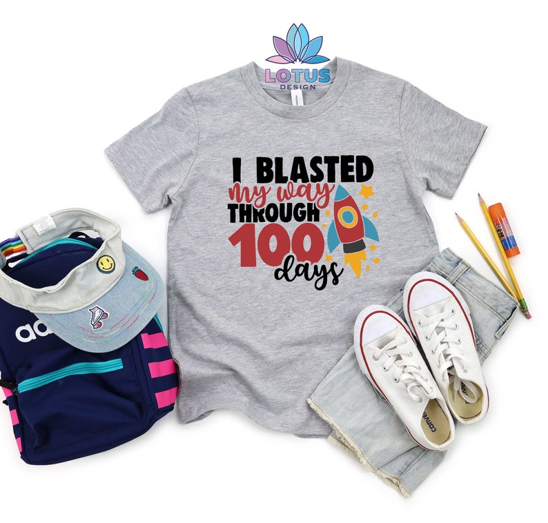 May include: A gray t-shirt with the text "I blasted my way through 100 days" and a red rocket ship graphic. The shirt is laid out on a white background with a pair of white sneakers, a denim skirt, and a backpack.