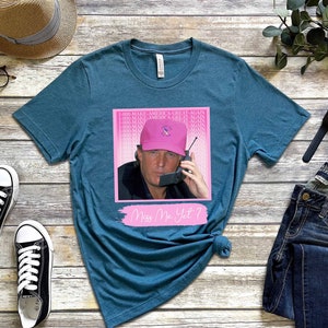 May include: Teal blue t-shirt with a pink and white graphic of a man wearing a pink hat and talking on a phone. The graphic has the text "I  MAKE AMERICA GREAT AGAIN" repeated multiple times. The text "Miss Me Yet?" is written in pink below the graphic.