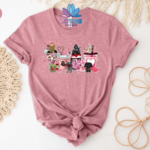 May include: A pink t-shirt with a graphic of Star Wars characters holding Valentine's Day themed drinks. The characters include Darth Vader, Yoda, Chewbacca, and others. The text "LOTUS DESIGN" is printed on the shirt.