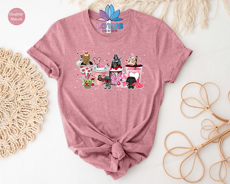 May include: A pink t-shirt with a graphic of Star Wars characters holding Valentine's Day themed drinks. The characters include Darth Vader, Yoda, Chewbacca, and others. The text "LOTUS DESIGN" is printed on the shirt.
