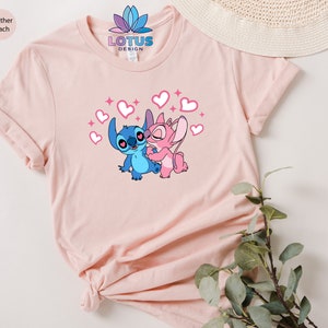 K&ouml;nnte beinhalten: Ein pinkfarbenes T-Shirt mit einem Motiv von Stitch und Angel aus dem Disney-Film Lilo & Stitch. Das Motiv zeigt Stitch und Angel, die sich umarmen, mit Herzen und Glitzer um sie herum. Der Text "LOTUS DESIGN" ist auf das Shirt gedruckt.
