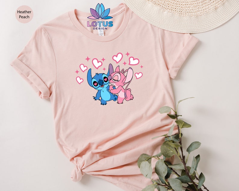 K&ouml;nnte beinhalten: Ein pinkfarbenes T-Shirt mit einem Motiv von Stitch und Angel aus dem Disney-Film Lilo & Stitch. Das Motiv zeigt Stitch und Angel, die sich umarmen, mit Herzen und Glitzer um sie herum. Der Text "LOTUS DESIGN" ist auf das Shirt gedruckt.
