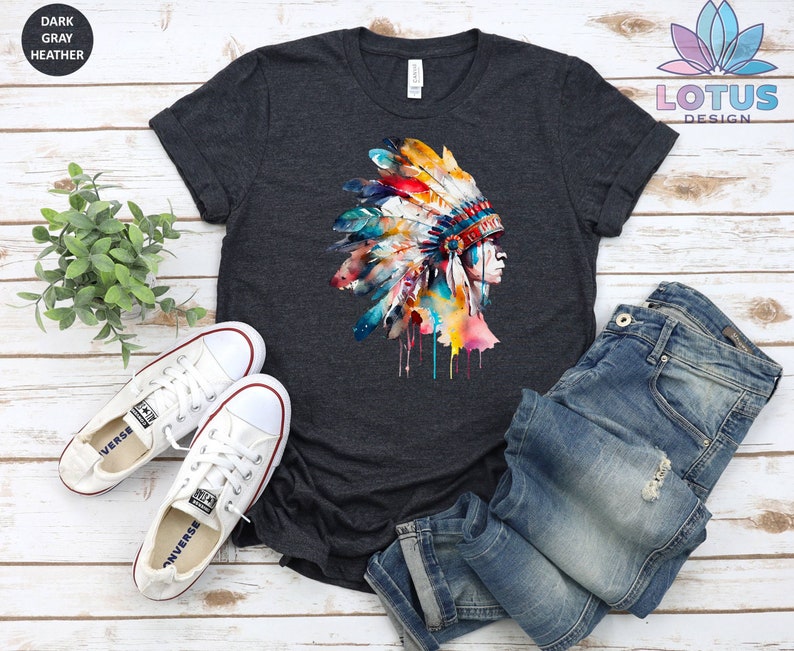 May include: A dark heather grey t-shirt with a watercolor illustration of a Native American headdress. The headdress is colorful with shades of blue, red, yellow, and green. The headdress is dripping with paint.