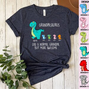 Op de afbeelding: Donkergrijs t-shirt met de tekst "GRANDPASAURUS" en een cartoon dinosaurus. Onder de tekst staan kleinere dinosaurussen met het woord "NAME" eronder. Het shirt zegt ook "LIKE A NORMAL GRANDPA, BUT MORE AWESOME". Een ontwerptabel met 12 dinosaurusontwerpen staat rechts.