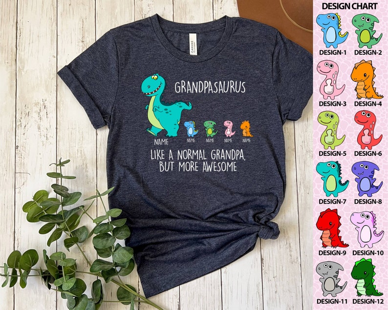 Op de afbeelding: Donkergrijs t-shirt met de tekst "GRANDPASAURUS" en een cartoon dinosaurus. Onder de tekst staan kleinere dinosaurussen met het woord "NAME" eronder. Het shirt zegt ook "LIKE A NORMAL GRANDPA, BUT MORE AWESOME". Een ontwerptabel met 12 dinosaurusontwerpen staat rechts.