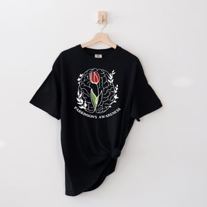 May include: Black t-shirt featuring a white outline of a brain with a red and green tulip inside. The text "Parkinson's Awareness" is written around the brain.
