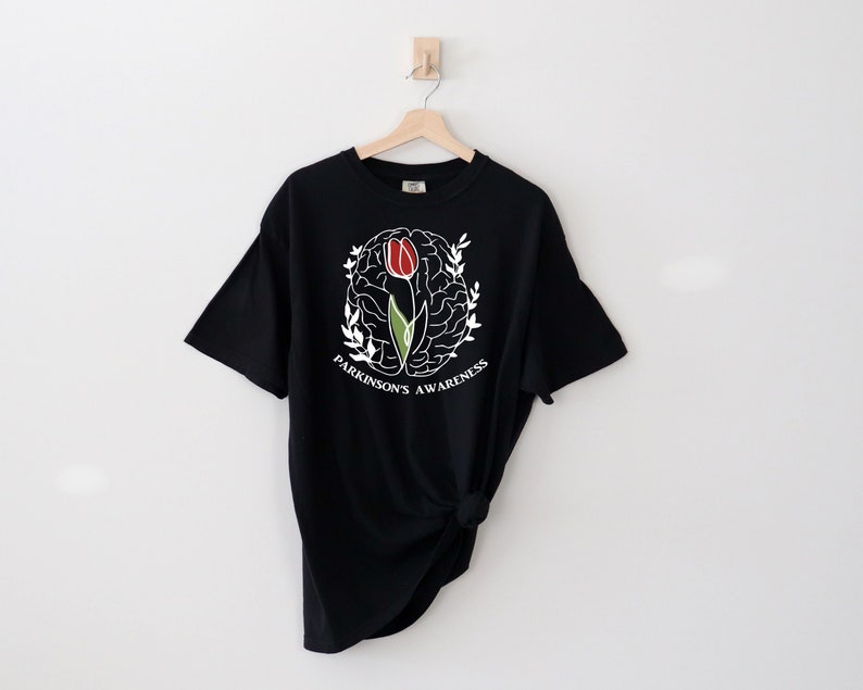 May include: Black t-shirt featuring a white outline of a brain with a red and green tulip inside. The text "Parkinson's Awareness" is written around the brain.