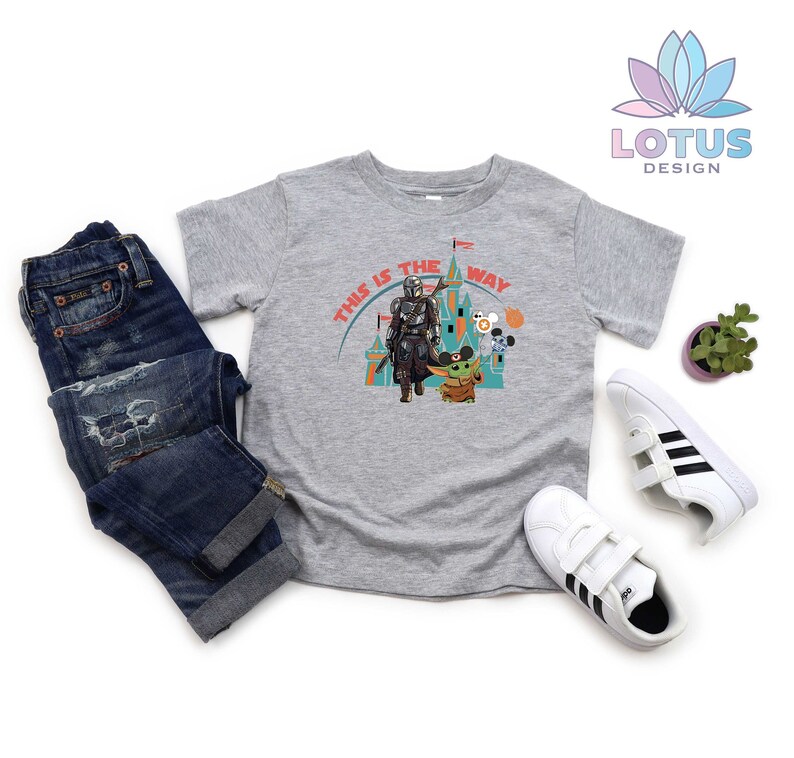 May include: A grey t-shirt featuring a graphic of a Mandalorian character and Grogu from Star Wars. The graphic includes the text "This is the way" and a castle-like structure.
