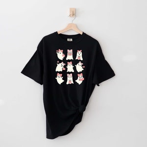 Może przedstawiać: Czarny T-shirt z grafiką dziewięciu białych duch&oacute;w w r&oacute;żowych kokardkach. Duchy są w r&oacute;żnych pozach, niekt&oacute;re trzymają serca, niekt&oacute;re machają, a niekt&oacute;re się uśmiechają.