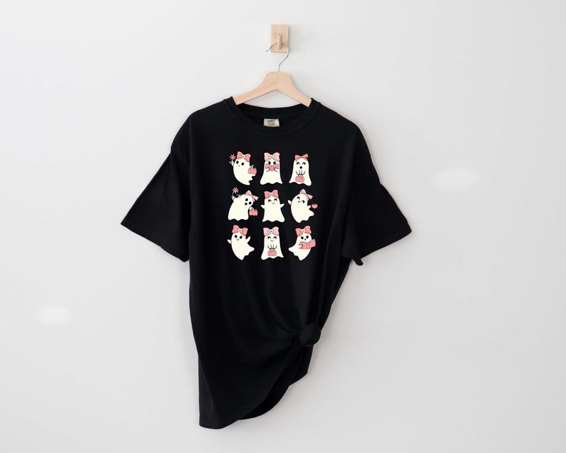 Może przedstawiać: Czarny T-shirt z grafiką dziewięciu białych duch&oacute;w w r&oacute;żowych kokardkach. Duchy są w r&oacute;żnych pozach, niekt&oacute;re trzymają serca, niekt&oacute;re machają, a niekt&oacute;re się uśmiechają.