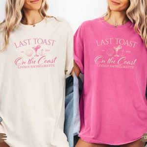 May include: Two t-shirts with the text "Last Toast On the Coast Linda's Bachelorette" in pink lettering. The shirts are in ivory and crunchberry colors.