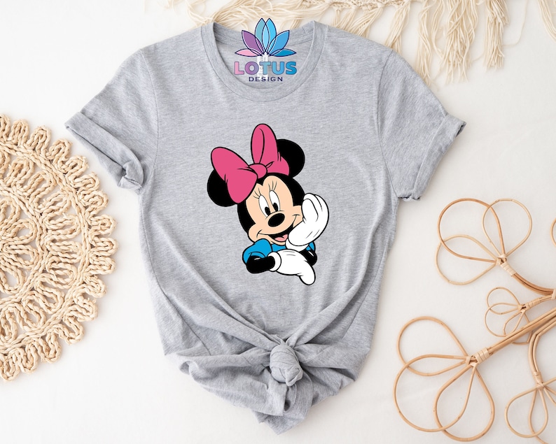 K&ouml;nnte beinhalten: Graues T-Shirt mit einem Motiv von Minnie Maus, die eine rosa Schleife und ein blaues Kleid tr&auml;gt. Das Shirt hat einen Knoten vorne.